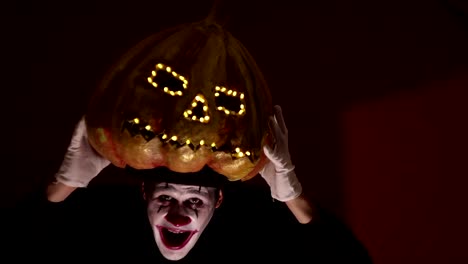 Scary-man-in-the-makeup-of-a-clown-holds-a-pumpkin-for-Halloween-and-puts-it-on-his-head.Terrible-clown-puts-on-a-mask-in-the-shape-of-a-pumpkin-on-his-head.-Terrible-clown-holds-in-his-hands-a-Jack-O-Lantern-with-luminous-eyes-and-a-mouth.