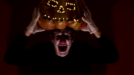 El-hombre-aterrador-en-la-composición-de-un-payaso-sostiene-una-calabaza-para-Halloween-y-se-la-pone-en-la-cabeza.-Un-payaso-terrible-se-pone-una-máscara-en-forma-de-calabaza-en-la-cabeza.-Terrible-payaso-sostiene-en-sus-manos-un-Jack-O-Lantern-con-ojos-