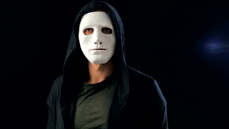 Anonymous-man-in-a-white-mask-covering-his-face-and-a-black-hood-turns-and-looks-at-the-camera.-Black-dark-background.-Concept-of-a-member-of-a-secret-organization