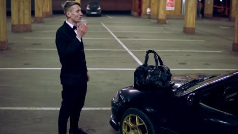 Young-man-in-a-black-suit-with-a-bow-tie-smoking-a-cigarette-by-the-black-car-in-the-parking.-Waiting-for-someone-and-holding-a-big-bag.-Being-nervous-and-impatient