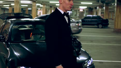 Young-man-in-a-black-suit-with-a-bow-tie-smoking-a-cigarette-sitting-at-the-bonnet-of-the-black-car-in-the-parking.-Waiting-for-someone.-Front-view.