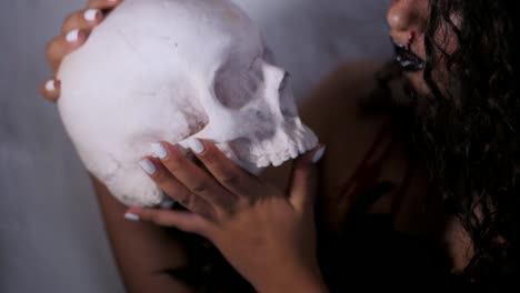 Scary-portrait-of-young-girl-with-Halloween-blood-makeup-holding-real-skull.-Beautiful-latin-woman-with-curly-hair-posing-in-studio.-Slow-motion