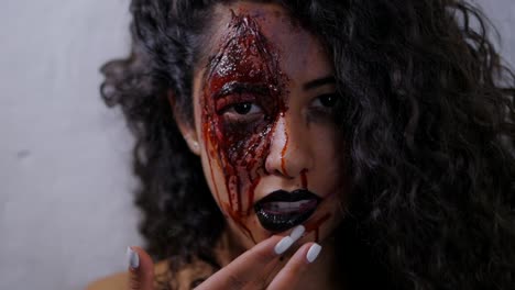 Scary-portrait-of-young-girl-with-Halloween-makeup-smears-blood-on-her-face.-Beautiful-latin-woman-with-curly-hair-looking-into-camera-in-studio.-Slow-motion