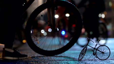 Lot-of-cyclists-ride-during-night-cycling-bike-parade-in-blur-by-illuminated-night-city-street-against-background-of-small-scale-model-of-bicycle.-Crowd-of-people-on-bike.-Bike-traffic.-Concept-sport-healthy-lifestyle.-Bright-shining-lights.-Low-angle