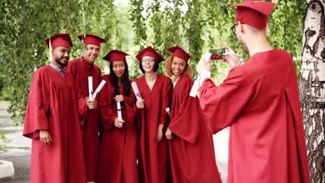 Alegres-estudiantes-graduados-están-de-pie-en-línea-al-aire-libre-y-joven-con-smartphone-es-disparos-mientras-que-los-graduados-se-ríen,-posando,-agitando-las-manos.