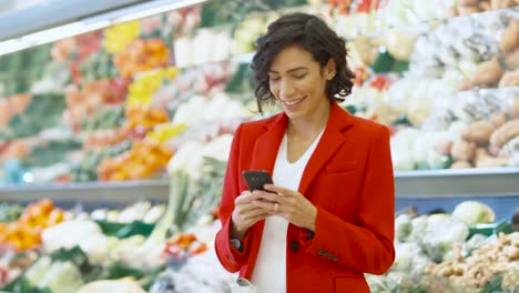 Im-Supermarkt:-schöne-junge-Frau-nutzt-Smartphone-stehen-im-Bereich-Frischprodukte-des-Ladens.-Im-Hintergrund-andere-Kunden-einkaufen.