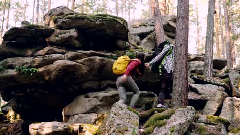 Adventurous-young-people-friends-are-trekking-in-forest-climbing-up-huge-rocks,-guy-is-helping-girl-giving-her-hand-during-hard-walk.-Adventures-and-friends-concept.