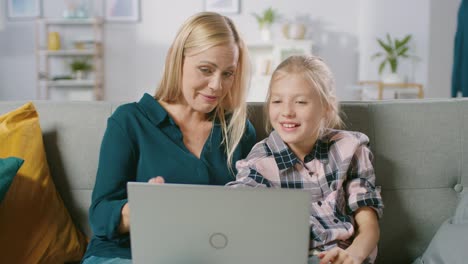 Hermosa-joven-madre-y-su-linda-pequeña-portátil-de-uso-hija-mientras-estaba-sentado-en-un-sofá-en-casa.-Familia-pasar-tiempo-junto-viendo-Videos-y-dibujos-animados-en-computadora.