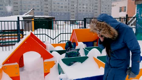 Mutter-ruft-auf-dem-Handy,-während-ihr-Sohn-auf-dem-Spielplatz-im-Winter-bei-Schneefall-geht.