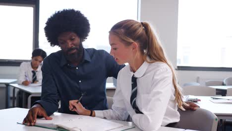 Tutor-de-secundaria-dando-estudiante-con-uniforme-enseñanza-uno-a-uno-en-el-escritorio-en-el-aula