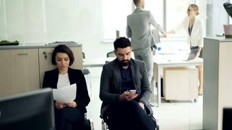 Young-people-man-and-woman-are-waiting-for-job-interview-in-office-while-manager-is-interviewing-another-candidate.-Girl-is-holding-cv,-guy-is-using-smartphone.