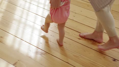 High-angle-view-of-cute-baby-girl-in-pink-bodysuit-making-her-first-steps-helped-by-caring-mom-at-home