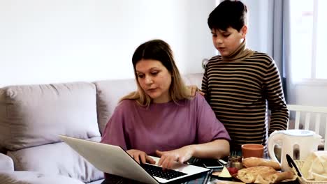 woman-working-at-laptop-and--sad-preteen-boy-standing-near-table