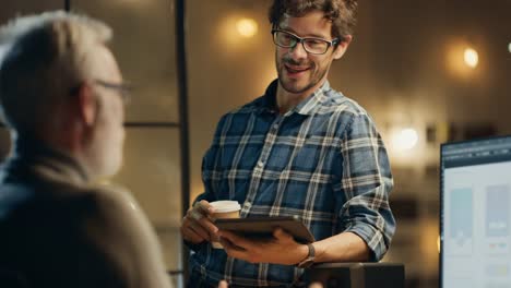 En-la-noche-creativo-de-mediana-edad-Smartphone-desarrollador-de-software-trabaja-en-un-ordenador-de-sobremesa-con-pantalla-que-muestra-el-diseño-de-la-aplicación,-tiene-conversación-con-el-especialista-más-joven-que-utiliza-la-tableta