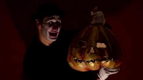 El-payaso-aterrador-tiene-una-máscara-de-calabaza-en-sus-manos.-Un-hombre-aterrador-con-maquillaje-de-payaso-sostiene-una-calabaza-para-Halloween.-Un-payaso-de-miedo-tiene-un-Jack-O-Lantern-en-sus-manos-con-ojos-luminosos-y-una-boca.