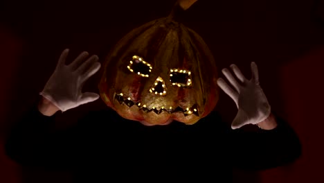 Hombre-aterrador-con-una-cabeza-de-calabaza-y-ojos-luminosos-mira-a-la-cámara.-Un-hombre-con-una-cabeza-de-calabaza-asusta-levantando-las-manos.-Víspera-de-Todos-los-Santos.