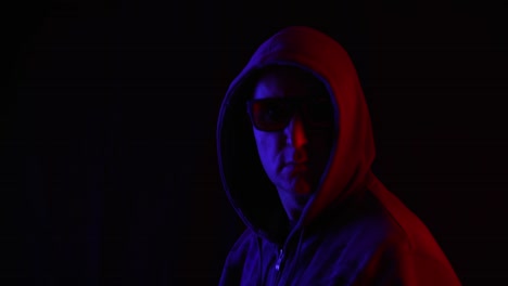 Portrait-adult-man-in-black-sunglasses-and-hoodie-turning-around-in-studio-on-black-background.-Man-in-hood-on-black-background-in-blue-and-red-lighting-looking-to-camera.