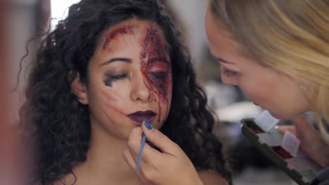 Make-up-artist-make-the-girl-halloween-make-upin-studio.Halloween-face-art.Woman-applies-on-professional-greasepaint-on-the-face-of-spanish-girl.War-paint-with-blood,-scars-and-wounds.Slow-motion