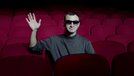 Single-man-sitting-in-comfortable-red-chairs-in-dark-cinema-theater-and-waving-hand-to-camera