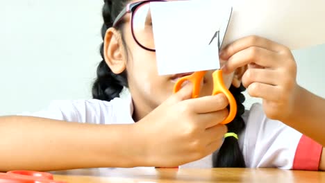 Niña-asiática-en-uniforme-de-estudiante-de-kindergarten-tailandés-usando-tijeras-para-cortar-el-papel-blanco-sobre-mesa-de-madera