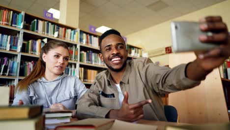 Africanos-merican-y-caucásico-se-divierten-sonriendo-y-tomando-fotos-selfie-en-cámara-del-smartphone-en-la-biblioteca-de-la-Universidad.-Los-estudiantes-alegres-tienen-descanso-mientras-preparan-para-examen