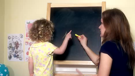 Teacher-woman-writing-numbers-on-chalk-black-board-for-little-child-girl