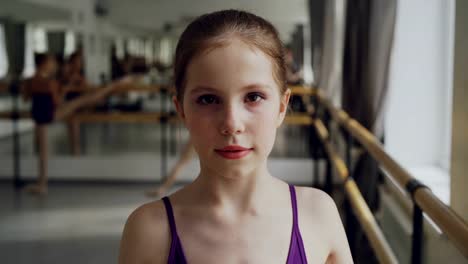 Portrait-of-young-girl-starting-ballet-dancer-with-make-up-looking-at-camera-and-smiling-during-ballet-class-in-spacious-light-dancehall.-Art-and-childhood-concept.