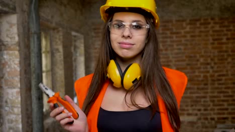 Young-beautiful-attractive-women-in-helmet,-earphones-and-glasses-dancing-with-tools,-holding-wrench,-screwdriver-and-pliers,-smiling,-female-builder,-brick-building-background