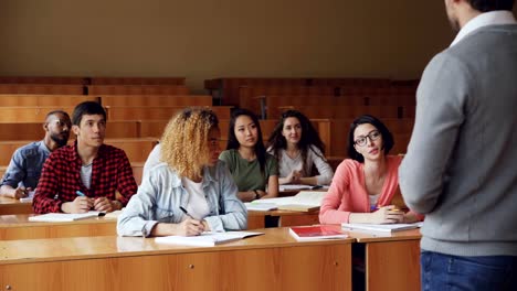 Hübsche-junge-Frau-fleißiger-Student-spricht-mit-High-School-Lehrer-am-Schreibtisch-sitzen,-während-andere-Schüler-Lächeln-hören-und-sind.-Moderne-Bildung-und-Jugend-Konzept.