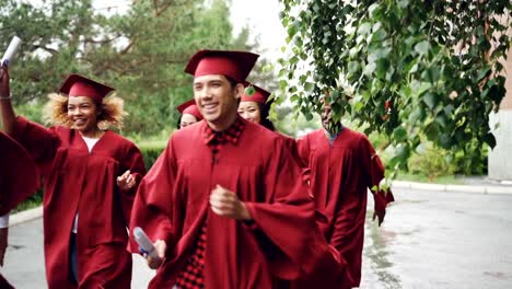 Slow-motion-of-happy-graduates-running-on-campus-waving-diplomas-and-smiling-wearing-red-gowns-and-hats.-Beautiful-trees-and-bushes-are-visible,-it-is-raining.