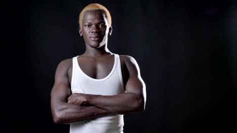 Young-african-blond-man-in-white-shirt-standing-with-crossed-hands-and-looking-at-camera,-confident-and-serious,-black-studio-background