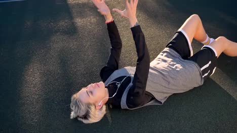 Charming-blonde-woman-lying-on-ground-and-playing-with-basketball,-listening-music-through-earphones,-beautiful-female-player