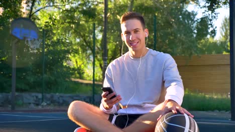 Handsome-happy-young-man-sitting-on-basketball-court-and-typing-on-phone,-holding-ball-and-listening-music,-smiling