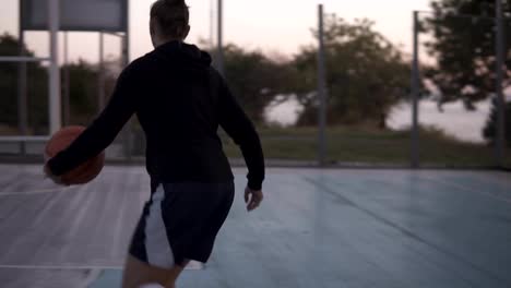 Backside-footage-of-a-young-basketball-female-player-jogging-with-ball-on-the-outdoors-court-and-throw-it-to-the-hoop