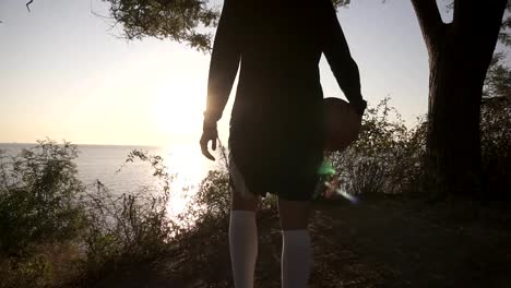 A-basketball-player-girl-comes-with-a-ball-in-her-hand,-coming-up-to-the-slope-with-trees-around.-Looks-at-the-sun-shining-over-the-sea.-Sea-view.-Backside-view