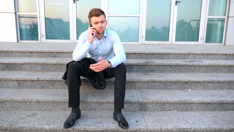 Dolly-shot-of-desperate-young-manager-talking-on-smart-phone-and-sitting-on-stairs-near-office-building.-Upset-sad-businessman-got-a-very-bad-news-on-cell-phone-and-crying-outdoor.-Close-up