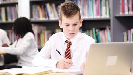 Estudiante-de-secundaria-de-hombre-usando-uniforme-trabajando-en-ordenador-portátil-en-la-biblioteca