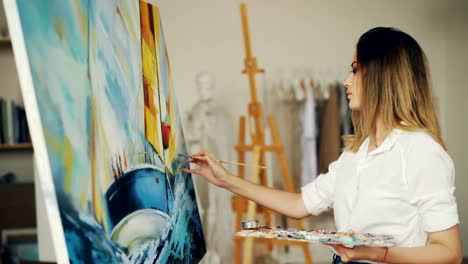 Blond-girl-painter-is-working-in-studio-depicting-sea-landscape-and-boat-on-canvas-using-brush-and-tempera-paints-looking-at-picture-and-enjoying-her-occupation.