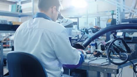 Electronics-Factory-Worker-in-White-Work-Coat-Inspects-a-Printed-Circuit-Board-Through-a-Digital-Microscope.-High-Tech-Factory-Facility.