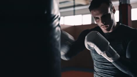 Ajuste-el-boxeador-entrenando-en-un-saco-de-boxeo-en-un-estudio-de-boxeo-interior