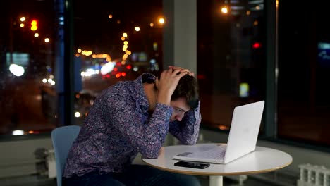 Der-Mann-Manager-sitzt-am-Laptop,-hielt-seinen-Kopf.-Erfahrungen.-Draußen-vor-dem-Fenster-die-nächtliche-Stadt.