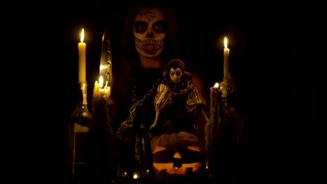 Bruja-de-Halloween-con-maquillaje-de-cráneo-hace-vudú-sostiene-cuchillo-y-wispering-hechizo-mágico-cadenas-de-calabaza-y-velas