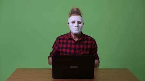 Hipster-man-wearing-mask-as-hacker-using-laptop-against-wooden-table