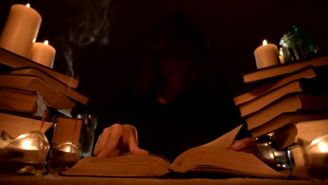 Una-niña-maga-de-primer-plano-mediana-en-una-capucha-en-una-habitación-oscura-a-la-luz-de-las-velas-y-en-busca-de-un-hechizo-que-da-vuelta-a-un-libro.-Llave-baja.