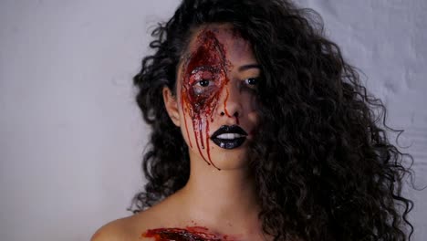 Scary-portrait-of-young-killer-girl-with-Halloween-blood-makeup.-Beautiful-latin-woman-with-curly-hair-looking-into-camera-in-studio.-Slow-motion