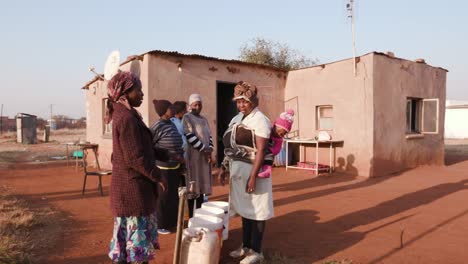 African-woman-collecting-water-from-a-tap-in-palstic-buckets,-while-others-stand-in-line-and-wait-their-turn