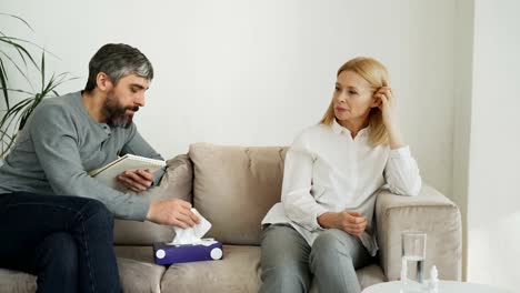 Crying-stressed-woman-talking-with-professional-male-psychologist-who-gives-paper-tissue-and-trying-to-calm-down-her-during-thearpy-session