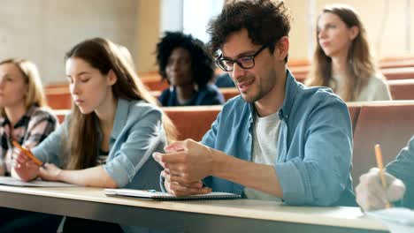 Hispanic-Mann-nutzt-Smartphone-während-einer-Vorlesung-im-Klassenzimmer.-Hörsaal-voller-Studenten.-Junge-Menschen-an-der-Universität.