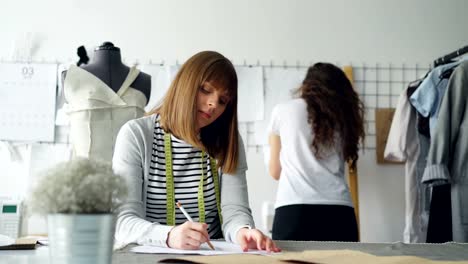 Young-clothing-designer-is-busy-drawing-sketch-with-pencil-while-her-colleague-is-watching-pictures-pinned-on-wall-in-design-studio.-Creative-thinking-concept.