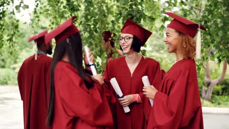 Compañeros-de-clase-están-hablando-y-riendo-después-de-la-ceremonia-de-graduación-con-diplomae-y-usar-vestidos-y-birretes,-chicas-compartiendo-recuerdos-y-expresando-esperanzas.